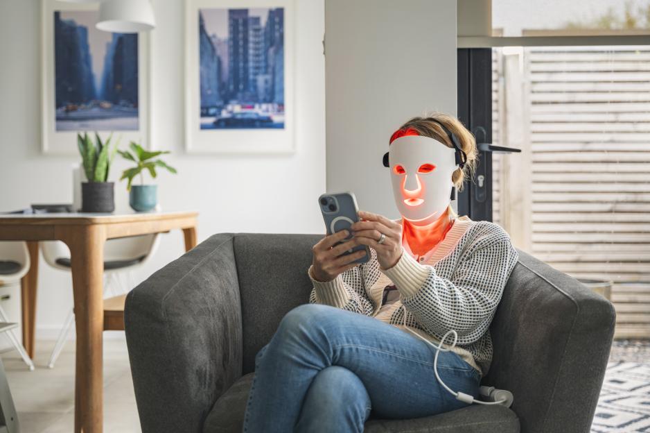 Woman using a red light therapy mask while relaxing on a couch and using her phone.