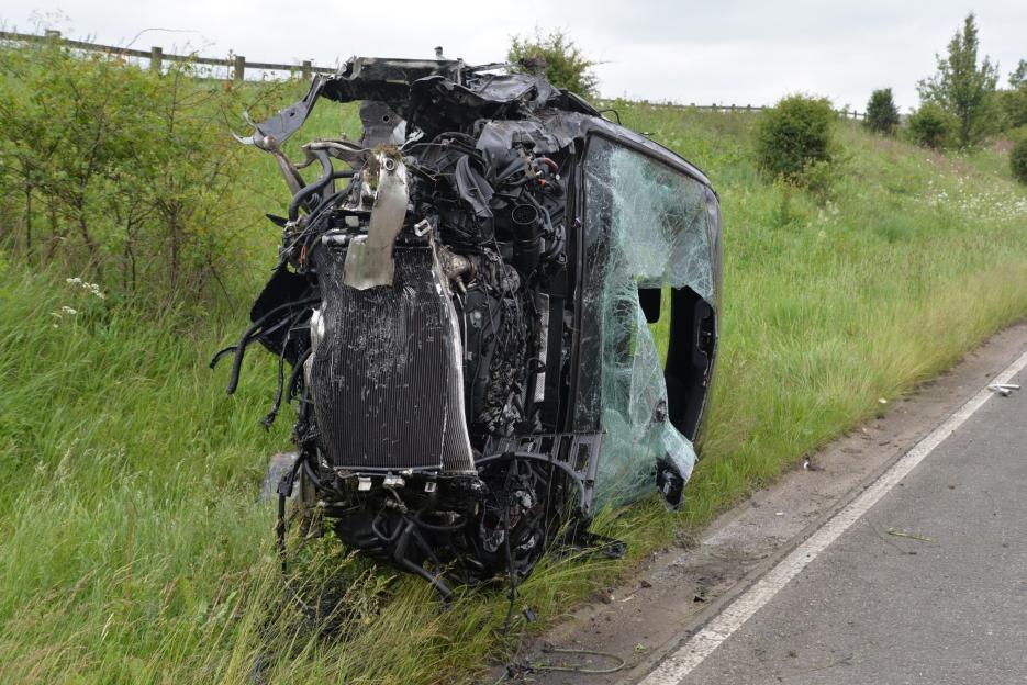 A severely damaged black Audi Q5 is at the side of a road, with its front end completely destroyed and windshield shattered.