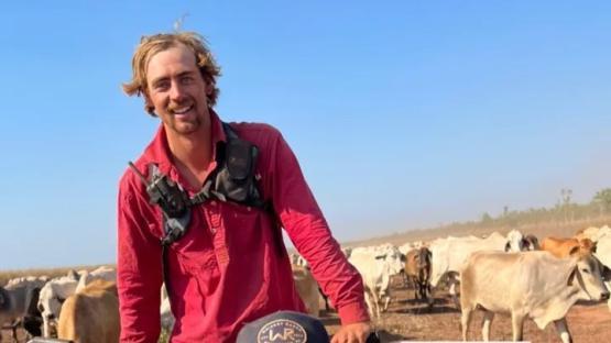 A smiling man in a red shirt stands in front of a herd of cattle.