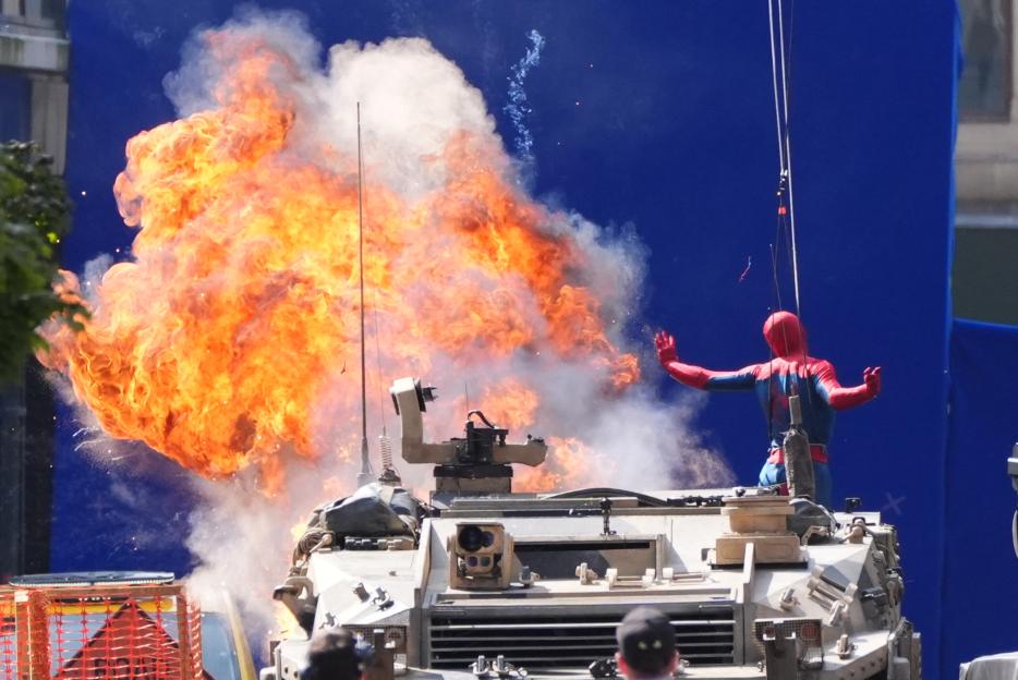 Spider-Man in costume on a military vehicle with an explosion behind him during filming.