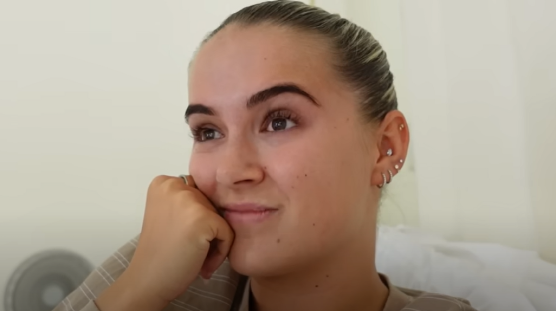 A young woman with dark hair pulled back, wearing a striped shirt, looks to the left with a slight smile, resting her chin on her hand.