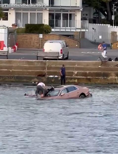A car mostly submerged in water, with a person on top and another person in the water next to it.