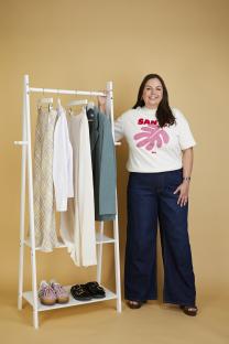 Woman standing next to clothing rack with various clothing items and shoes.