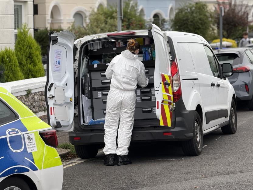 A person in a white forensic suit and black boot covers stands at the back of a white van with its rear doors open, organizing items. A police vehicle is partially visible on the left.