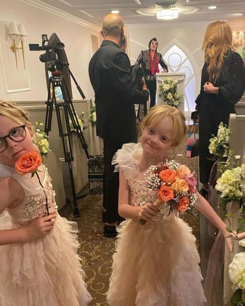 Two young girls, dressed in light-colored ruffled dresses, hold flowers in the foreground of a wedding chapel where an Elvis impersonator is officiating.