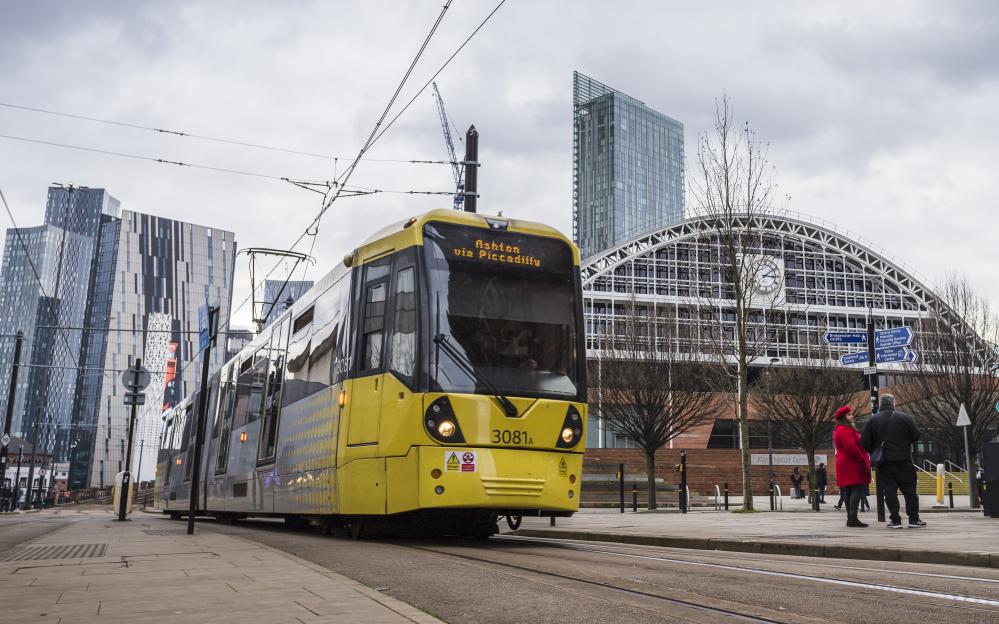 Yellow Metrolink tram arriving at a station, with tall buildings and a large arched building in the background.