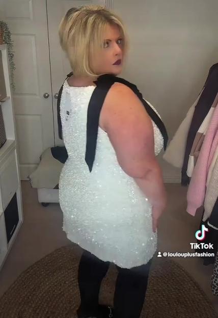 A plus-size person models a white sparkly mini dress with black bow shoulder straps.
