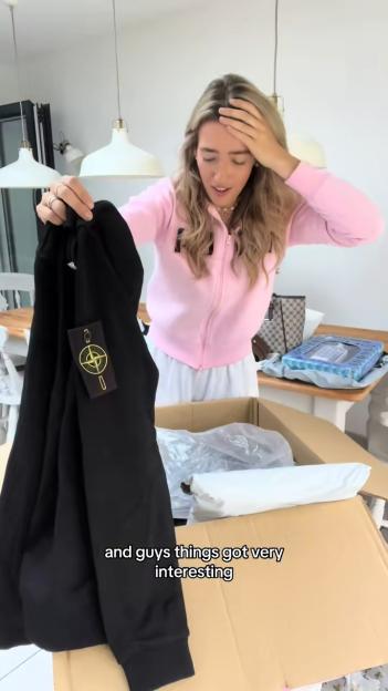 A young woman in a pink top unboxes a package, revealing a black long-sleeved shirt with a Stone Island badge.
