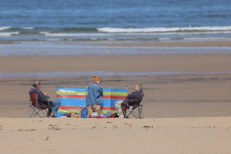 Three people sitting in chairs on a beach behind a windbreak.