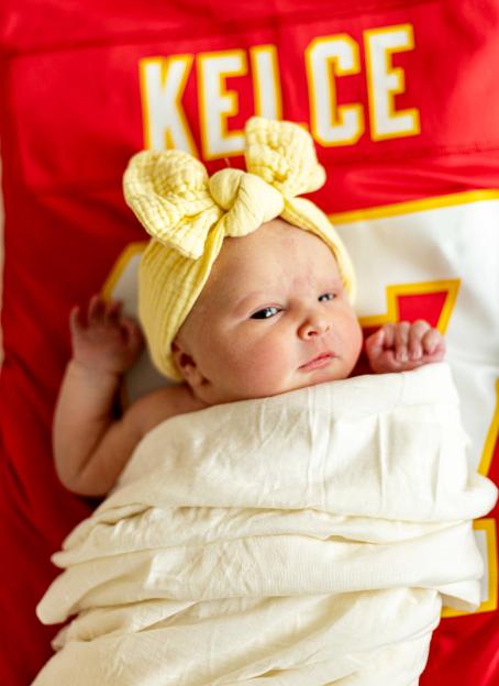 Newborn baby girl lying on a red jersey, wearing a yellow headband.