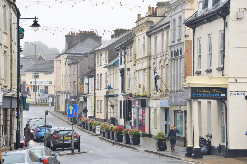 A street in Callington, Cornwall with cars parked along the side and a person walking on the sidewalk.