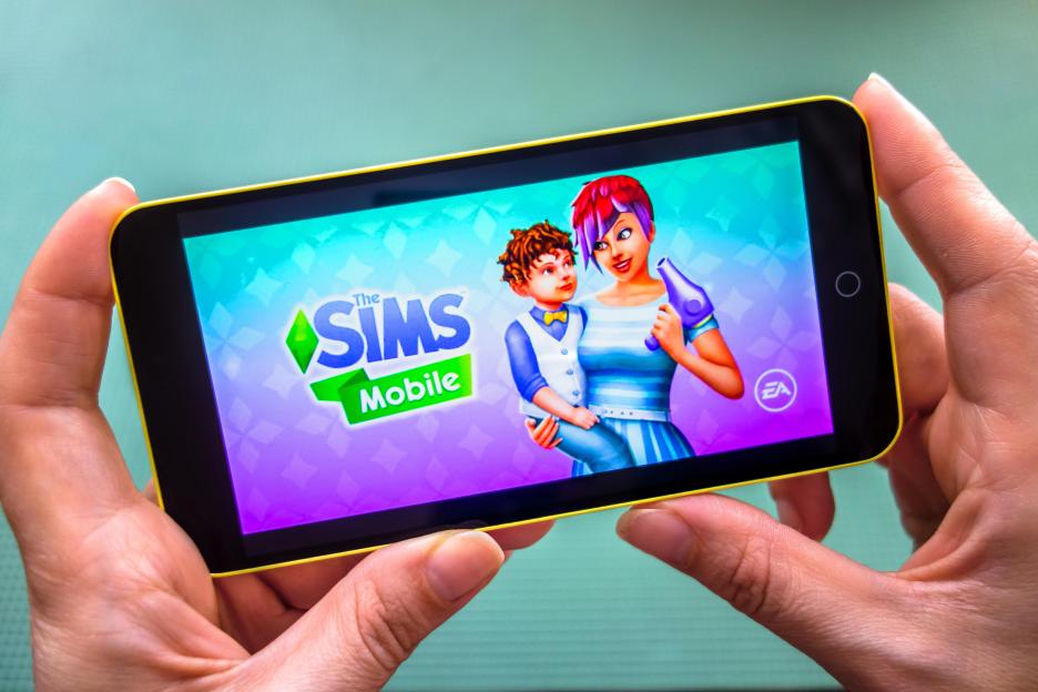 Close-up of hands holding a phone displaying "The Sims Mobile" game.