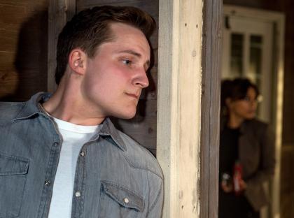 A man looking worried stands near a doorway while a woman stands in the background.