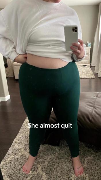 A person in a white top and green leggings stands with their midriff exposed, holding a phone to take a mirror selfie, with text overlay "She almost quit".