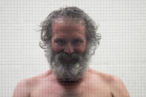 Shirtless man with long gray hair and beard behind glass.
