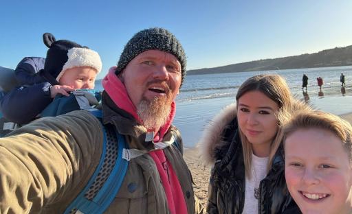 A man with a baby on his back and two teenagers beside him at the beach.