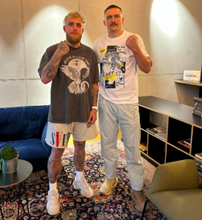 Two men standing together, one in a grey t-shirt and beige shorts, the other in a white t-shirt and light grey pants.