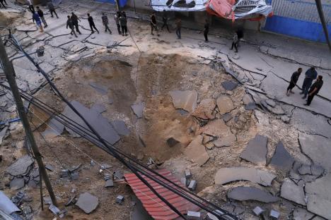 Aftermath of an airstrike, showing a large crater in a road and people surveying the damage.