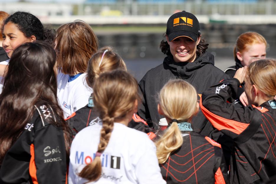 A woman wearing a black racing hat and jacket smiling at a group of young girls.