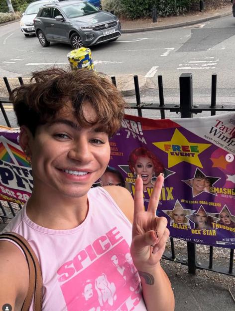 A person in a pink "SPICE" shirt making a peace sign in front of a poster for an event with drag queens.