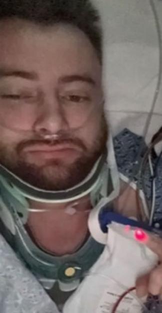 Mitchell Deakin in a hospital bed with a neck brace and nasal cannula after a skydiving accident.