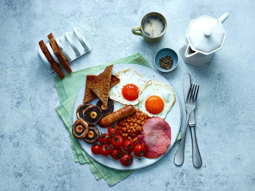 A breakfast plate featuring fried eggs, sausage, bacon, baked beans, roasted tomatoes, mushrooms, and toast, next to a cup of coffee.