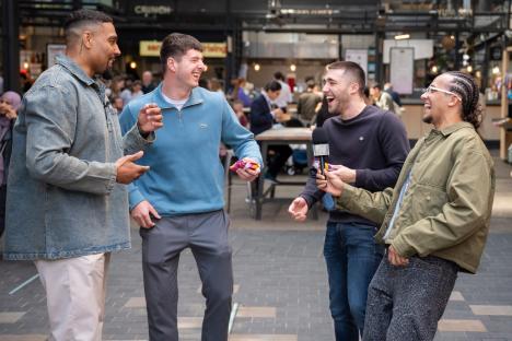 Four men laughing and holding snacks and microphones outdoors.