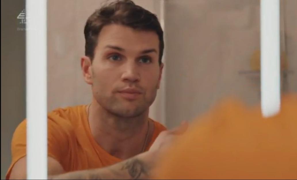 Man in orange shirt looking at his reflection in a mirror.