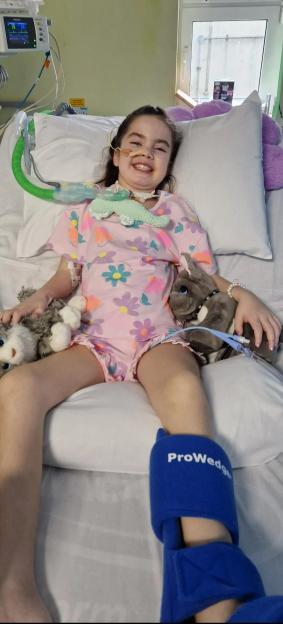 Ivy Amos, 9, paralysed from the neck down, smiles in her hospital bed with medical equipment and stuffed animals.