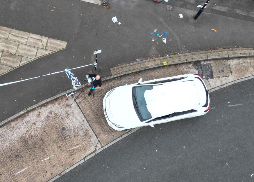 Overhead view of a crime scene with police tape, a white car, and a police officer.