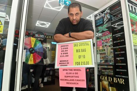 A vape shop owner stands with arms crossed in front of a sign advertising vape juice and a raffle to support Macmillan Cancer Research.