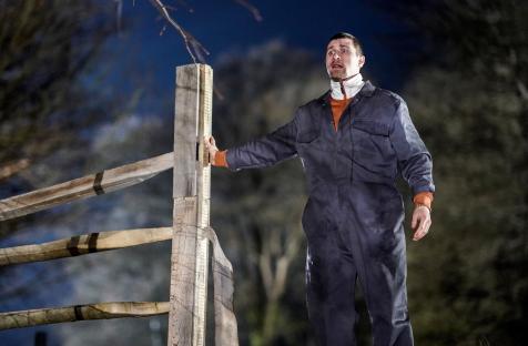 A man in a jumpsuit looking over a damaged wooden fence at night.