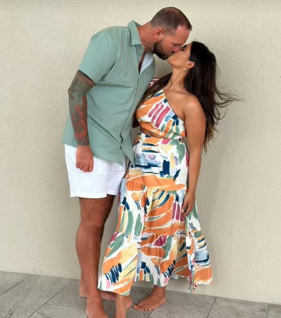 A tall man in a green shirt and white shorts kisses a woman in a colorful abstract patterned dress.