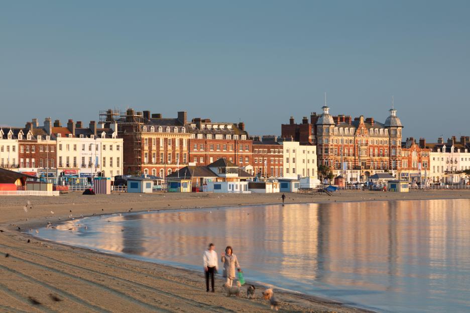 Two people walk their dogs on Weymouth Beach at dawn, with colorful buildings and beach huts in the background.