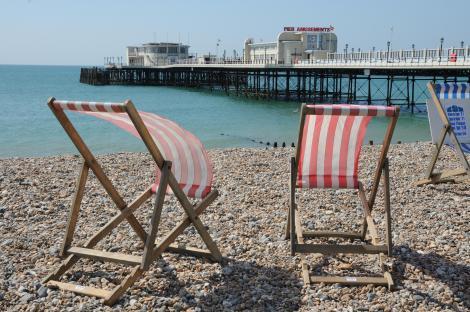 Wooden deck chairs on a pebble beach with a pier in the background.