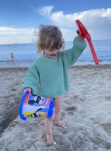 Toddler on beach with Mickey Mouse bucket and red sand toy.