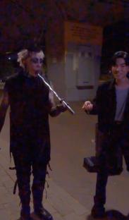 A person in dark clothing holding a sword-like object near another person.