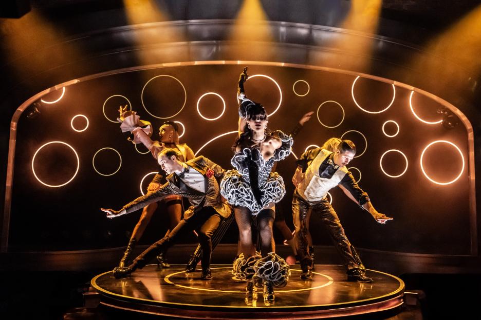 Five circus performers on a round stage with glowing orange circles in the background.