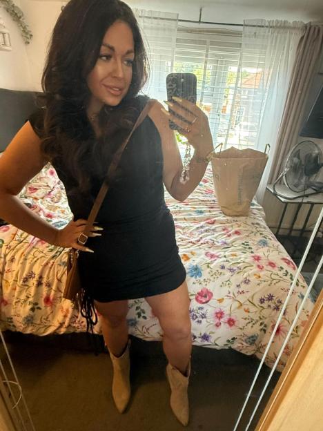 Woman in black dress and beige boots taking a mirror selfie.
