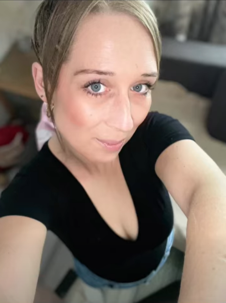 A woman with blonde hair, blue eyes, and a black top, poses for a selfie.