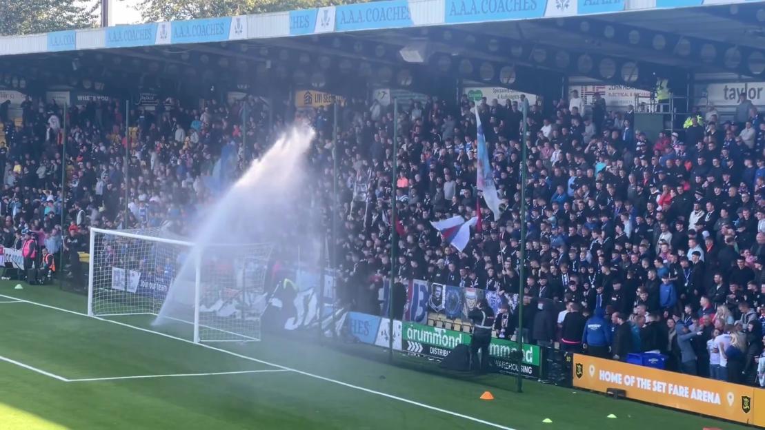 A soccer game with fans in the stands and a sprinkler spraying water across the goal.