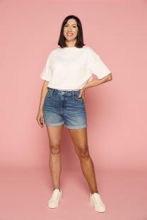 Woman in white shirt and denim shorts smiling against pink background.
