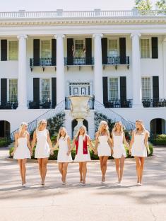 Seven women in white dresses walking in front of a large white house.
