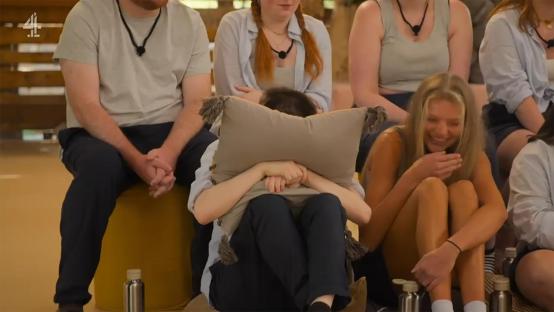 A person hides their face behind a pillow while others laugh.