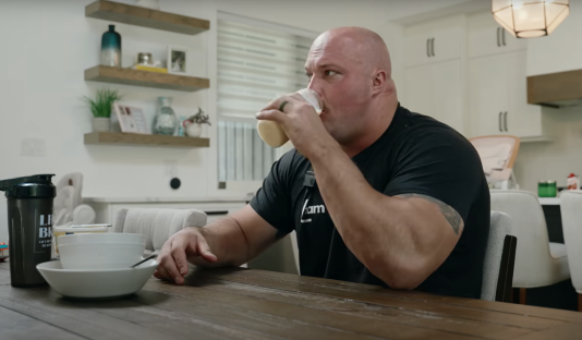 A large man sits at a table drinking from a cup.