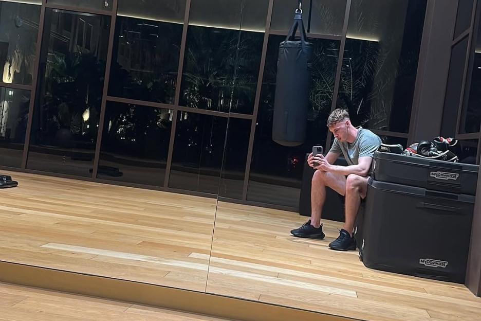 Cole Palmer sitting in a gym with a punching bag, looking at his phone.
