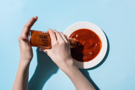 Hands pouring tomato sauce from a bottle onto a plate.