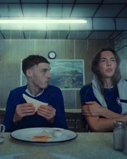 Two people wearing Chelsea football jerseys eat toast in a diner.