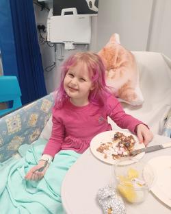 Girl with pink hair eating a treat in a hospital bed.