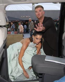 Woman in a light green gown getting into a car, with a man holding up a hand to stop people.